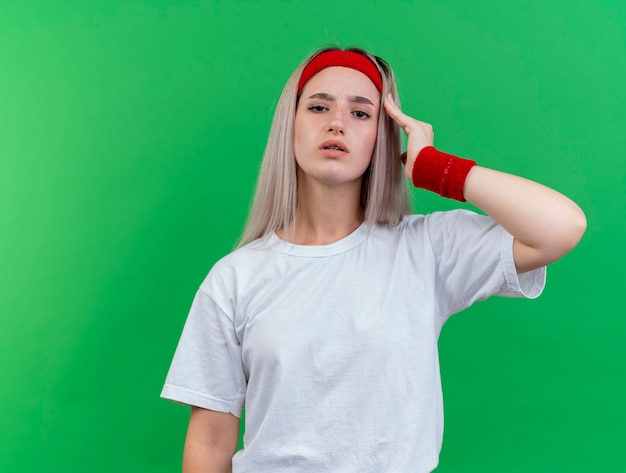 Fiduciosa giovane donna sportiva con le parentesi graffe che indossa la fascia e braccialetti mette la mano sul tempio isolato sulla parete verde