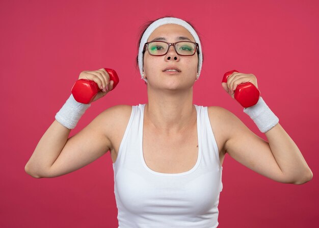 Fiduciosa giovane donna sportiva in occhiali ottici che indossa fascia e braccialetti tiene manubri isolati sulla parete rosa