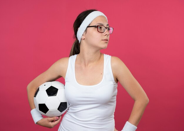 Fiduciosa giovane donna sportiva in occhiali ottici che indossa la fascia e braccialetti tiene la palla e guarda al lato isolato sulla parete rosa