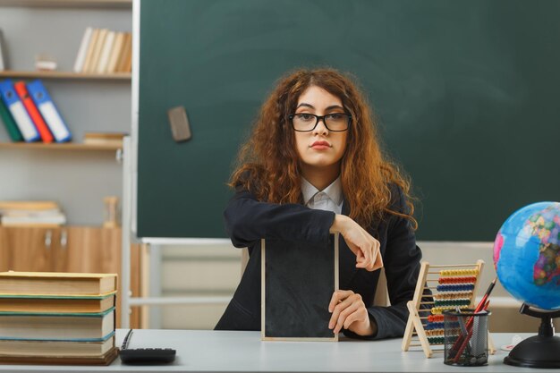fiduciosa giovane insegnante femminile che indossa occhiali con in mano una mini lavagna seduta alla scrivania con strumenti scolastici in classe