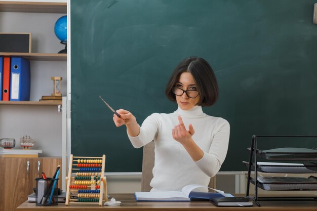 fiduciosa giovane insegnante femminile che indossa occhiali punta a lato con il puntatore seduto alla scrivania con gli strumenti scolastici in classe