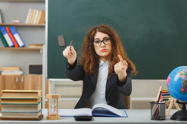 fiduciosa giovane insegnante femminile che indossa occhiali punta alla telecamera con il puntatore seduto alla scrivania con strumenti scolastici in classe
