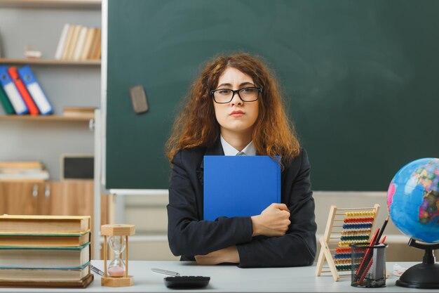 fiduciosa giovane insegnante femminile con gli occhiali che tiene il libro seduto alla scrivania con gli strumenti della scuola in classe