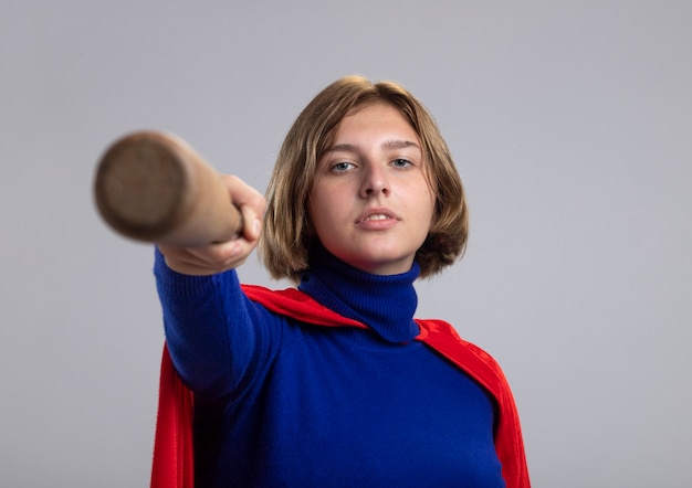 Fiduciosa giovane ragazza bionda del supereroe in mantello rosso che allunga la mazza da baseball verso la macchina fotografica che guarda l'obbiettivo isolato su priorità bassa bianca con lo spazio della copia