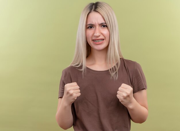 Fiduciosa giovane ragazza bionda in parentesi graffe dentali con i pugni serrati su uno spazio verde isolato con spazio di copia