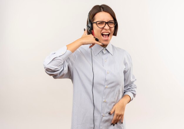 Fiduciosa giovane ragazza call center con gli occhiali e auricolare facendo segno di chiamata isolato su bianco con copia spazio