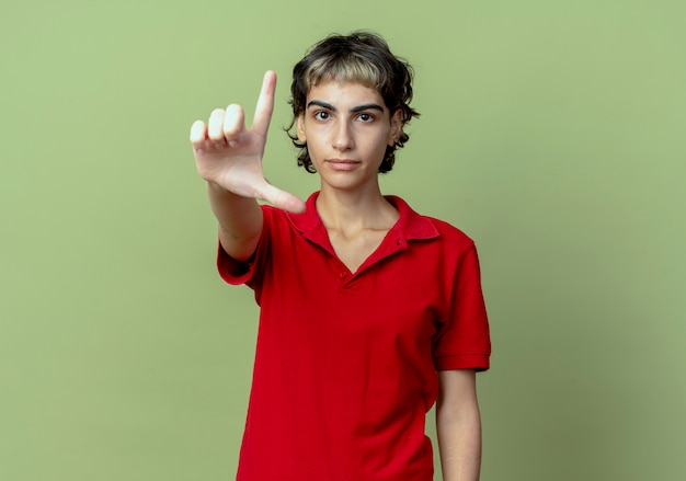 Fiduciosa giovane ragazza caucasica con taglio di capelli pixie facendo il gesto perdente in telecamera isolata su sfondo verde oliva con lo spazio della copia