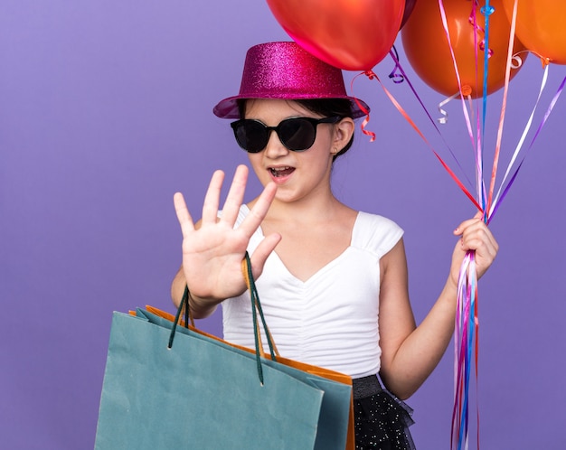 fiduciosa giovane ragazza caucasica in occhiali da sole con cappello da festa viola che tiene palloncini di elio e borse della spesa che gesturing il segnale di stop isolato sulla parete viola con spazio di copia