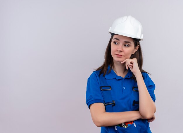 Fiduciosa giovane ragazza costruttore con casco di sicurezza bianco e uniforme blu mette la mano sul mento e guarda al lato su sfondo bianco isolato con spazio di copia