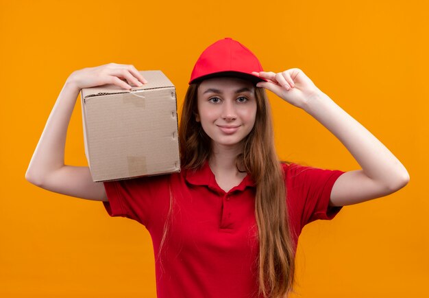 Fiduciosa giovane ragazza di consegna in rosso uniforme tenendo la scatola sulla spalla e mettendo la mano sul cappuccio su uno spazio arancione isolato