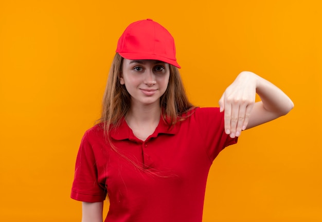 Fiduciosa giovane ragazza di consegna in uniforme rossa che punta con la mano sullo spazio arancione isolato