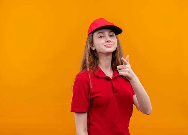 Fiduciosa giovane ragazza di consegna in uniforme rossa che ti fa gesto sullo spazio arancione isolato con lo spazio della copia