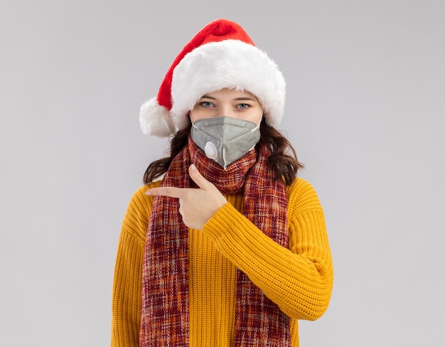 Fiduciosa giovane ragazza slava con il cappello della santa e con la sciarpa intorno al collo che indossa la mascherina medica che indica al lato