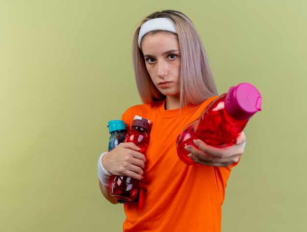 Fiduciosa giovane ragazza sportiva caucasica con bretelle che indossa fascia e braccialetti tiene le bottiglie d'acqua guardando la fotocamera