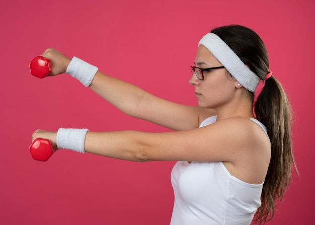 Fiduciosa giovane ragazza sportiva in occhiali ottici che indossa fascia e braccialetti sta lateralmente tenendo i manubri