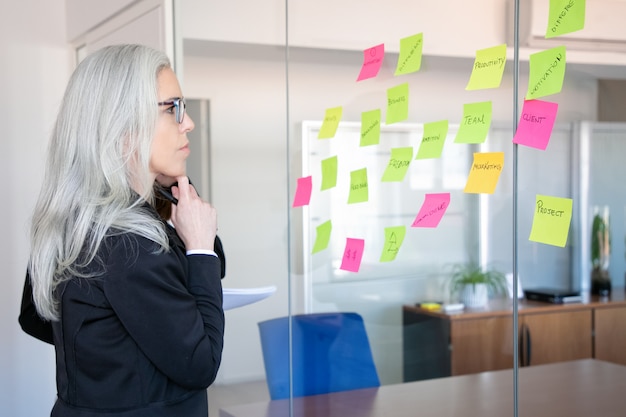 Fiduciosa imprenditrice concentrata guardando gli adesivi sulla parete di vetro. Lavoratore femminile dai capelli grigi concentrato che pensa alle note per la strategia di progetto. Marketing, affari e concetto di gestione