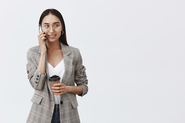 Fiduciosa imprenditrice in giacca e occhiali, guardando a destra con espressione intrigata e gioiosa mentre utilizza lo smartphone e beve caffè