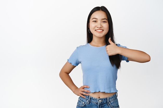Fiduciosa ragazza asiatica sorridente mostra i pollici in su dare l'approvazione dire sì e lode in piedi in maglietta su sfondo bianco