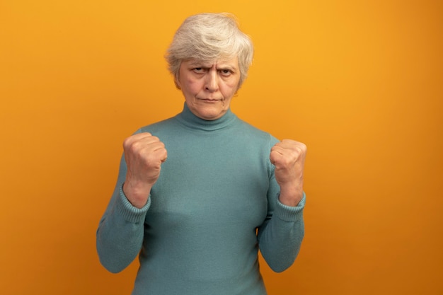 Fiduciosa vecchia donna che indossa un maglione a collo alto blu guardando i pugni serrati davanti facendo essere un gesto forte isolato sulla parete arancione con spazio di copia