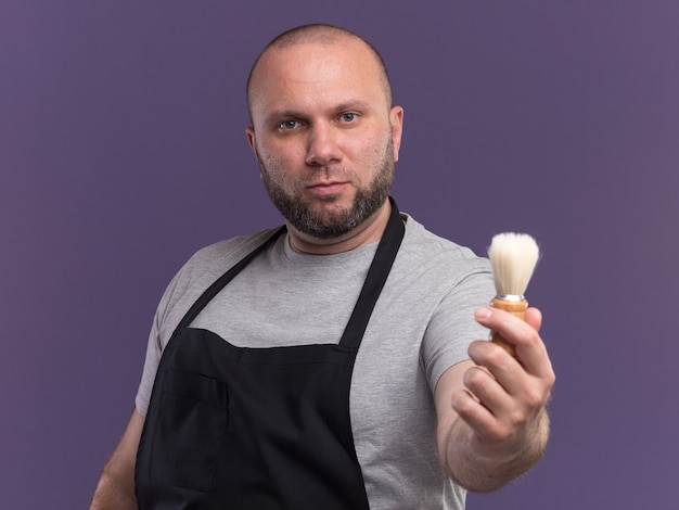 Fiducioso barbiere maschio di mezza età in uniforme che porge il pennello da barba alla telecamera isolata sul muro viola