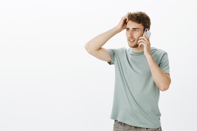 Fiducioso bel ragazzo biondo, guardando a sinistra e toccando i capelli tenendo lo smartphone vicino all'orecchio e parlando, discutendo di qualcosa e pensando a quale risposta