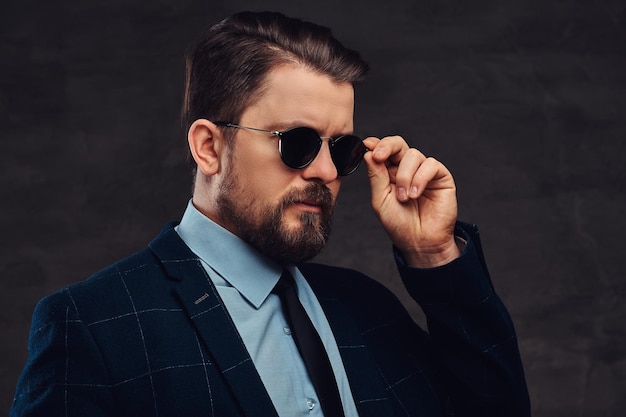 Fiducioso bell'uomo di mezza età alla moda con barba e acconciatura vestito con un elegante abito formale e occhiali da sole su uno sfondo scuro strutturato in studio.