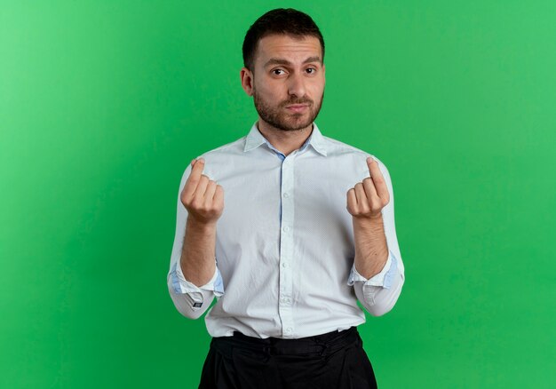 Fiducioso bell'uomo gesti il segno della mano dei soldi con due mani isolate sulla parete verde
