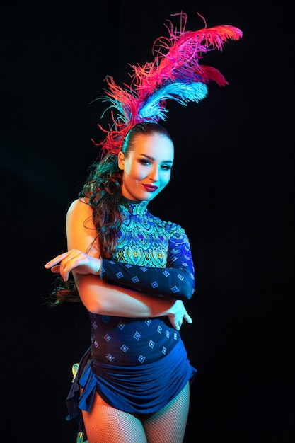 Fiducioso. Bella giovane donna in carnevale, elegante costume in maschera con piume sul muro nero in luce al neon. Copyspace per annuncio. Celebrazione delle feste, balli, moda. Tempo festivo, festa.