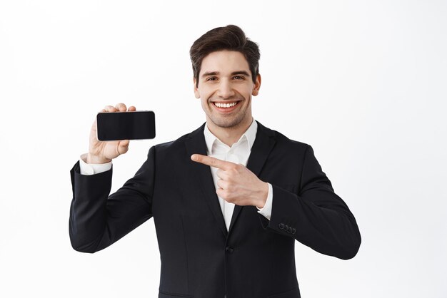 Fiducioso capo impiegato maschio che punta allo schermo orizzontale del telefono e sorridente dimostra il sito Web o l'applicazione in piedi in tuta su sfondo bianco