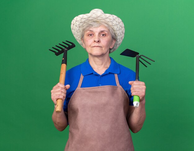 Fiducioso donna anziana giardiniere che indossa cappello da giardinaggio che tiene rastrello e rastrello zappa