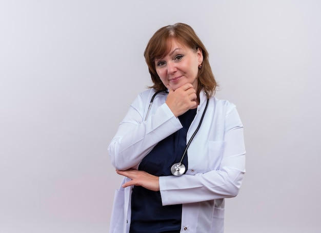 Fiducioso donna di mezza età medico indossando abito medico e stetoscopio mettendo la mano sul mento sulla parete bianca isolata con lo spazio della copia