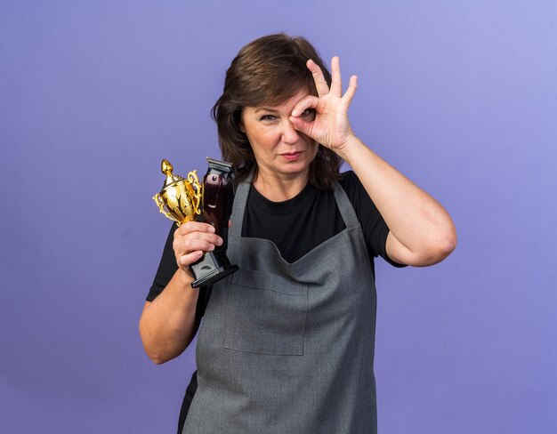 Fiducioso femmina adulta barbiere in uniforme tenendo il tagliacapelli e la tazza del vincitore guardando la parte anteriore attraverso le dita isolate sulla parete viola con spazio di copia