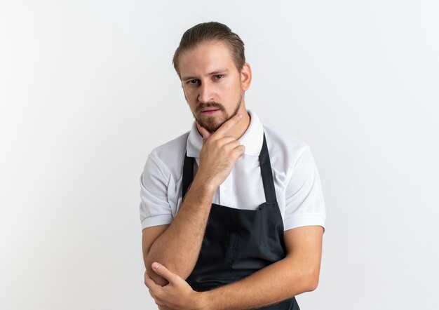 Fiducioso giovane barbiere bello che indossa l'uniforme mettendo la mano sul mento e un altro sul gomito isolato su bianco con lo spazio della copia