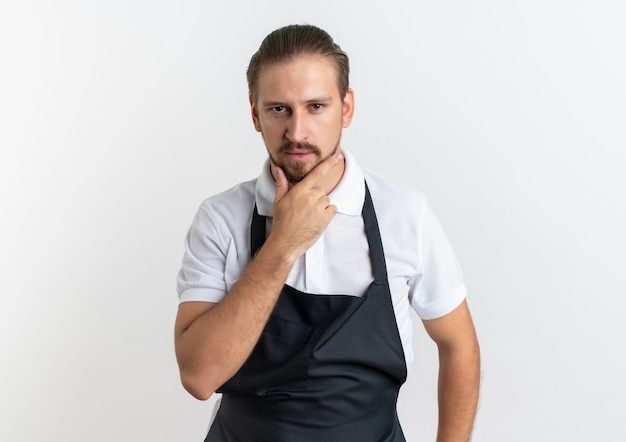 Fiducioso giovane barbiere bello che indossa l'uniforme mettendo la mano sul mento isolato su bianco con lo spazio della copia