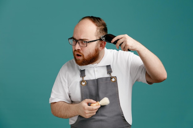 Fiducioso giovane barbiere che indossa l'uniforme e gli occhiali con in mano un pennello da barba tagliando i propri capelli con un rifinitore per capelli mentre guarda la fotocamera isolata su sfondo blu