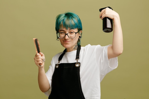 Fiducioso giovane barbiere che indossa occhiali uniforme che tiene pettini guardando la fotocamera applicando spray per capelli sui propri capelli isolati su sfondo verde oliva