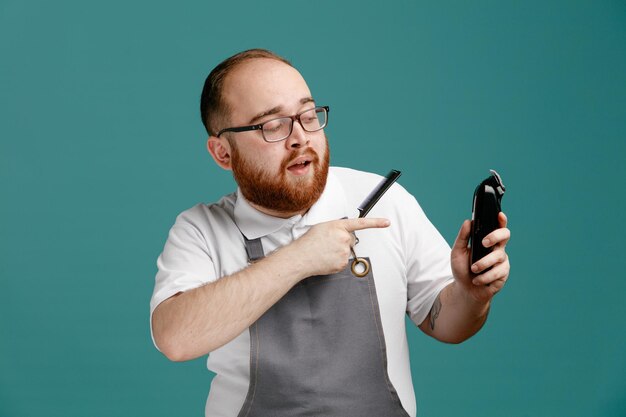 Fiducioso giovane barbiere che indossa uniforme e occhiali con pettine teaser e rifinitore per capelli guardando e indicando il rifinitore per capelli isolato su sfondo blu