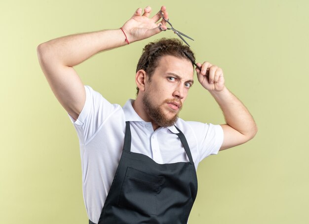 Fiducioso giovane barbiere maschio che indossa l'uniforme tagliando i capelli con le forbici isolate sulla parete verde oliva