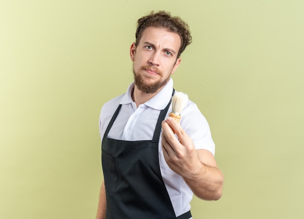 Fiducioso giovane barbiere maschio che indossa l'uniforme tenendo fuori il pennello da barba isolato su parete verde oliva