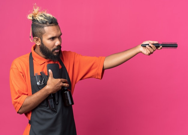 Fiducioso giovane barbiere maschio che indossa l'uniforme tenendo pettini e lacca per capelli guardando e puntando con il pettine a lato isolato su parete rosa