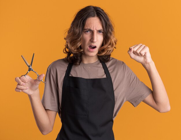 Fiducioso giovane bel barbiere che indossa l'uniforme tenendo le forbici facendo un gesto forte