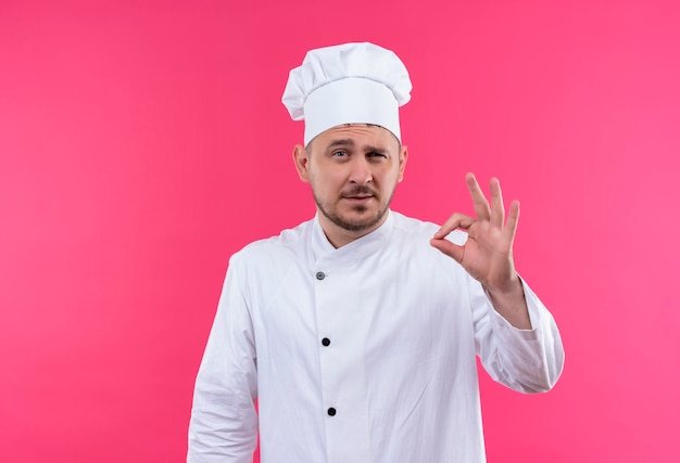 Fiducioso giovane bel cuoco in uniforme da chef che fa segno ok isolato su parete rosa