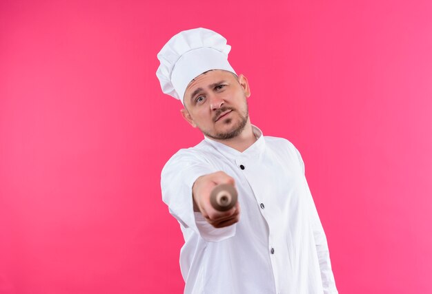 Fiducioso giovane bel cuoco in uniforme da chef che punta con il mattarello isolato sulla parete rosa