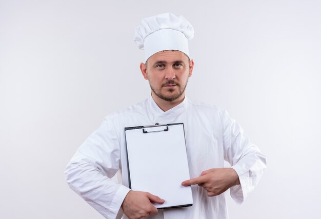 Fiducioso giovane bel cuoco in uniforme da chef che tiene e indica la lavagna per appunti isolata sul muro bianco