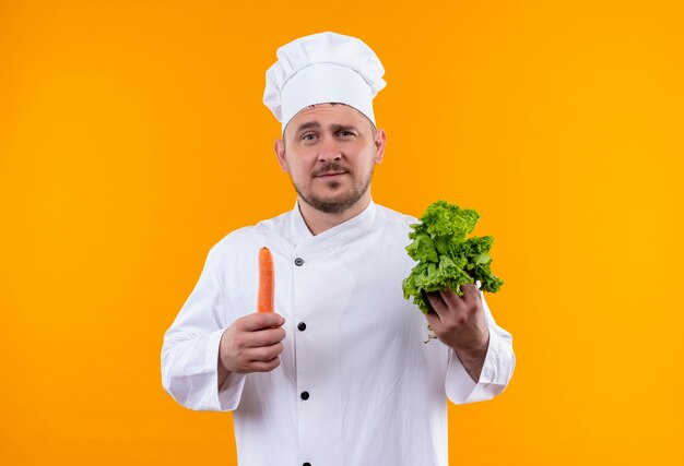 Fiducioso giovane bel cuoco in uniforme da chef che tiene lattuga e carota isolate sulla parete arancione