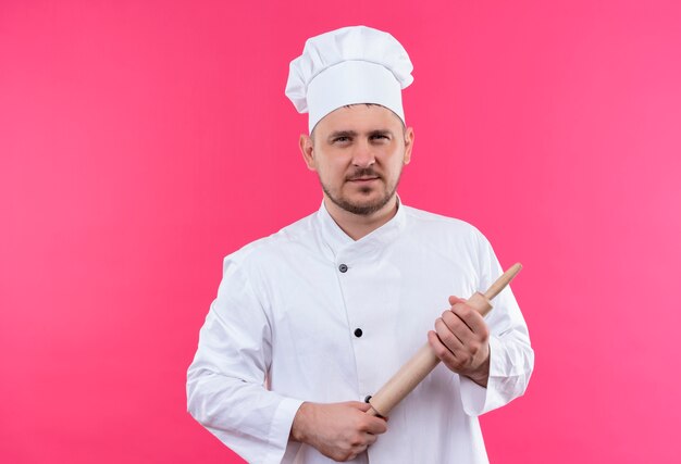 Fiducioso giovane bel cuoco in uniforme da chef che tiene mattarello isolato su parete rosa