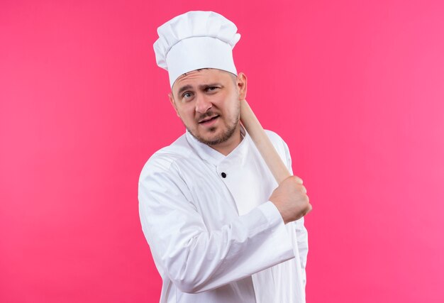 Fiducioso giovane bel cuoco in uniforme da chef che tiene mattarello isolato su parete rosa