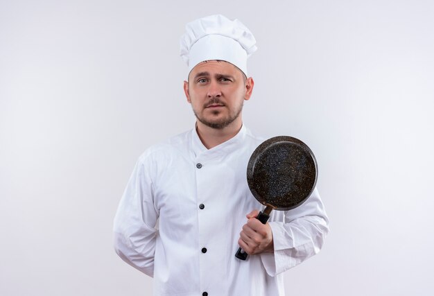 Fiducioso giovane bel cuoco in uniforme da chef che tiene padella isolata sul muro bianco
