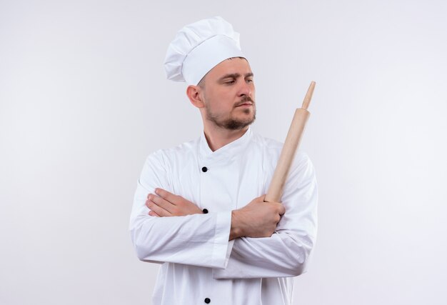 Fiducioso giovane bel cuoco in uniforme da chef in piedi con postura chiusa e tenendo il mattarello e guardando il lato isolato sul muro bianco