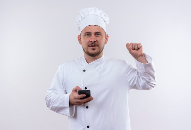 Fiducioso giovane bel cuoco in uniforme da chef tenendo il telefono cellulare e alzando il pugno isolato sul muro bianco